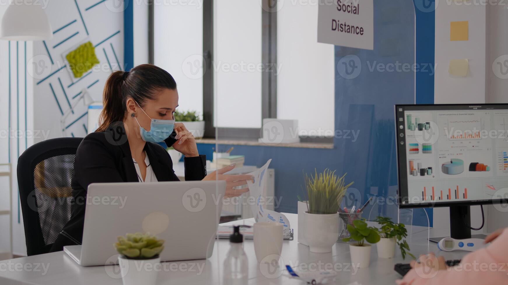 Woman with protective face mask working on laptop computer in company office checking statistics while talking on phone.Team workers respecting social distancing to avoid infection with covid19 virus photo