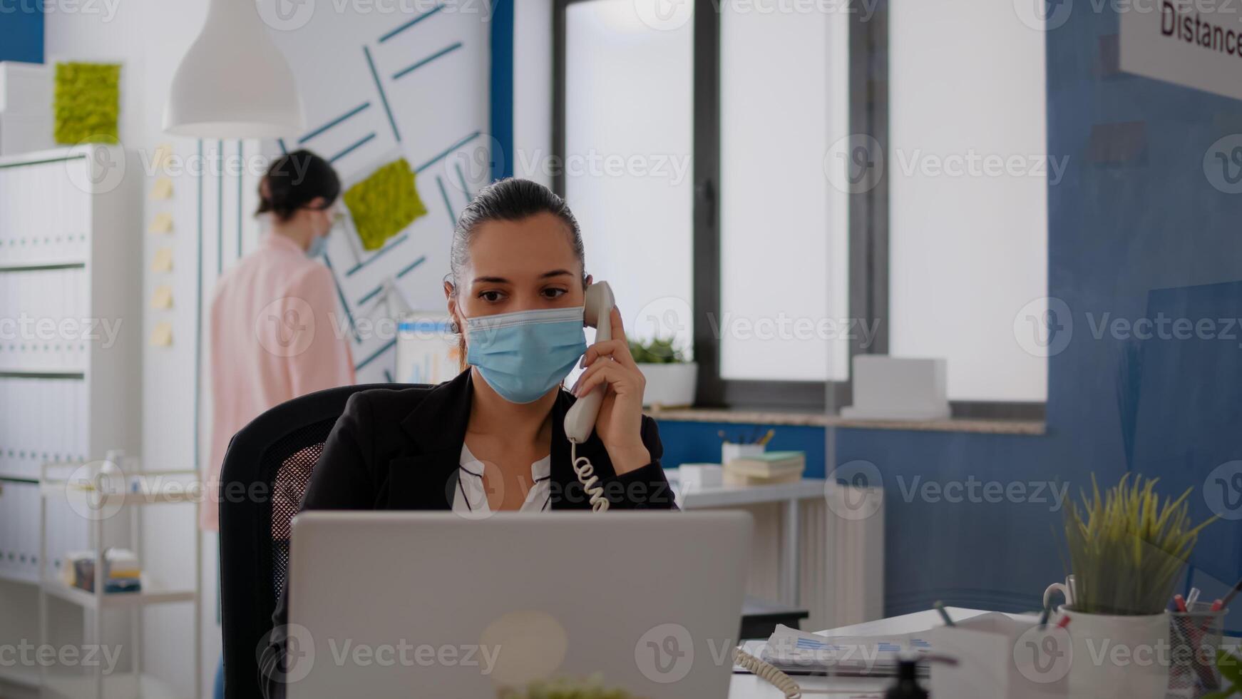 Entrepreneur with protective face mask discussing on landline while sitting at startup office desk in front of computer. Caucasian female working at business meeting during coronavirus global pandemic photo
