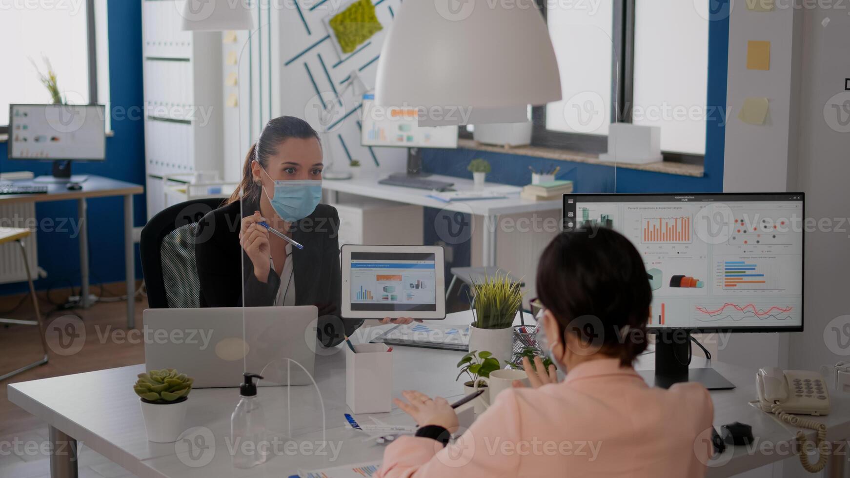 Businesswoman with face mask analyzing statistics with her collegue using digital tablet computer. Team working in new normal office respecting social distance to prevent infection with covid19 photo