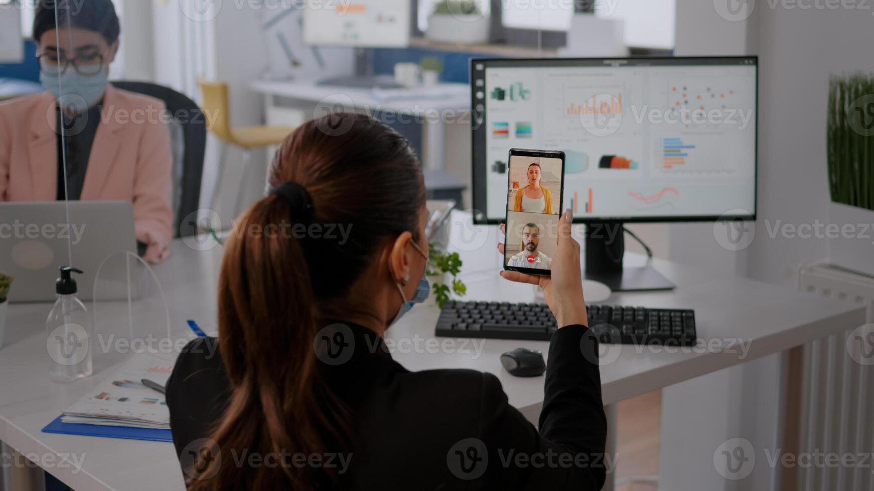Executive business woman wearing protective face mask using phone for online videocall conference with remotely team. Coworkers working in background respecting social distance photo