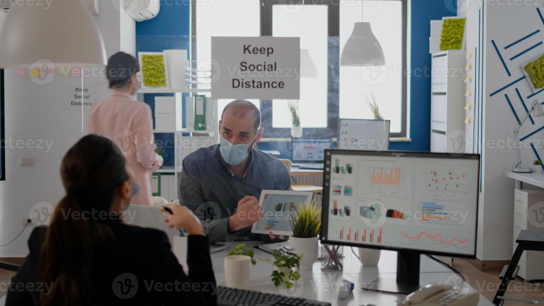 Group of business coworkers with face masks analyzing graphs using digital tablet sitting in new normal workplace. Colleague working in background mantain social distancing photo