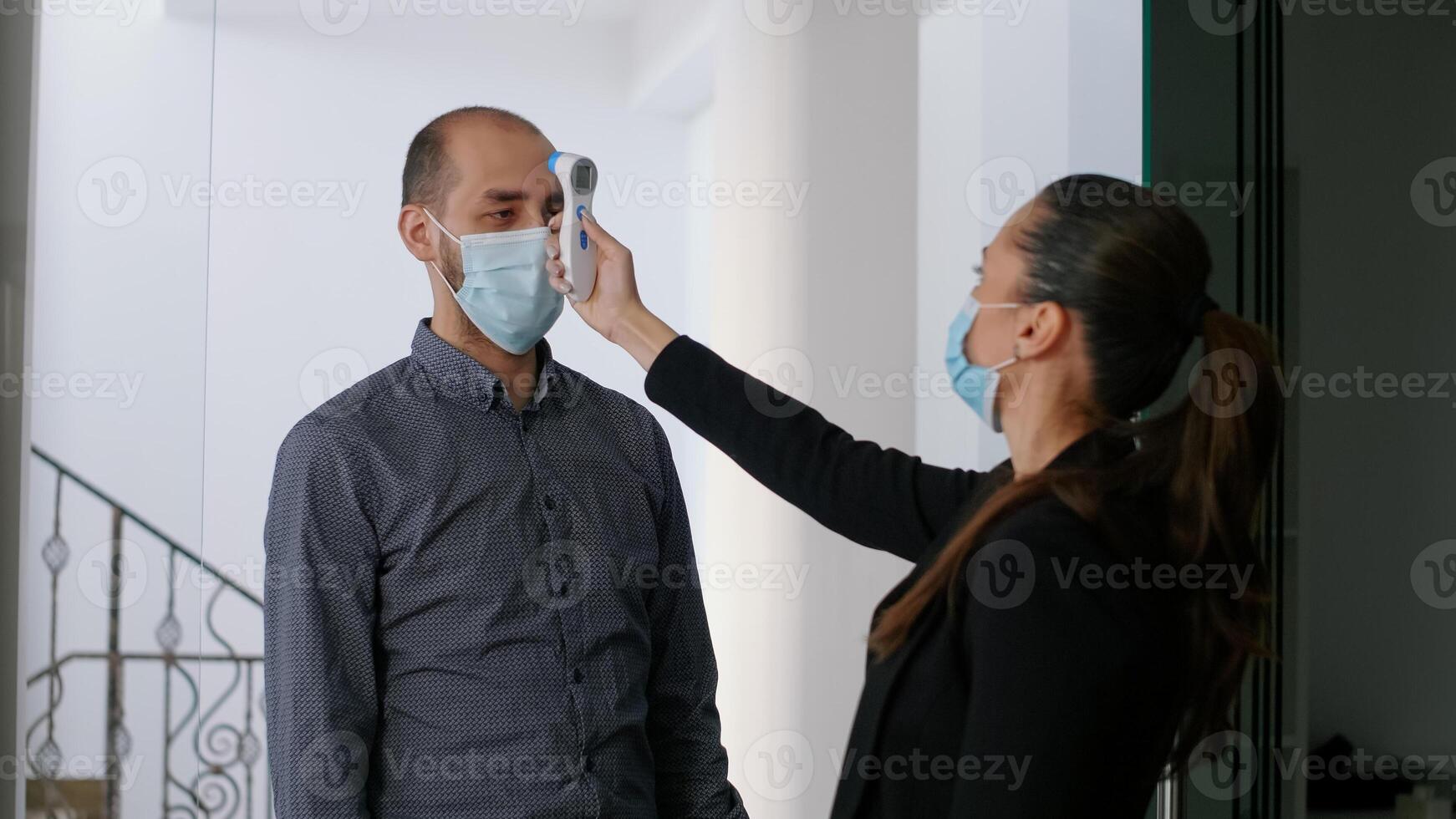 Caucasian female wearing face mask checking workers temperature using infrared thermometer. Team respecting social distance in new normal company office to avoid infection with virus disease photo
