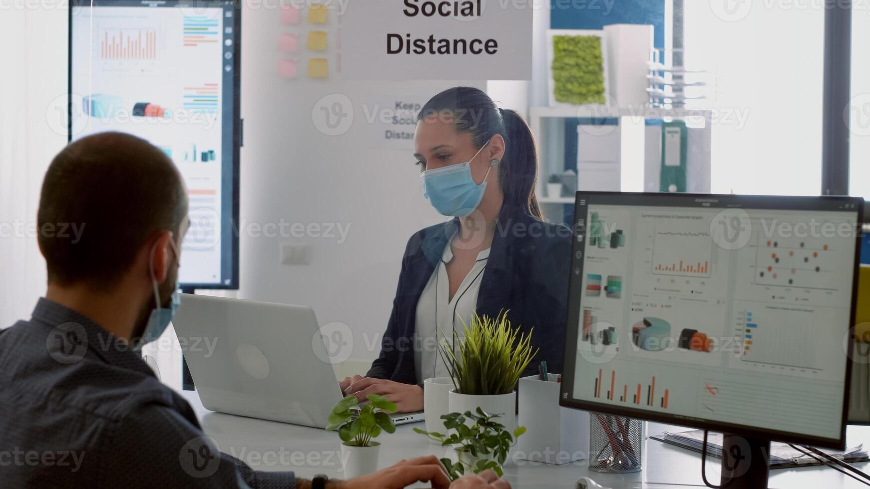Businessman looking at financial graphs on computer display while talking with coworker sitting in company office. Colleagues with face masks keeping social distancing to prevent covid19 disease photo