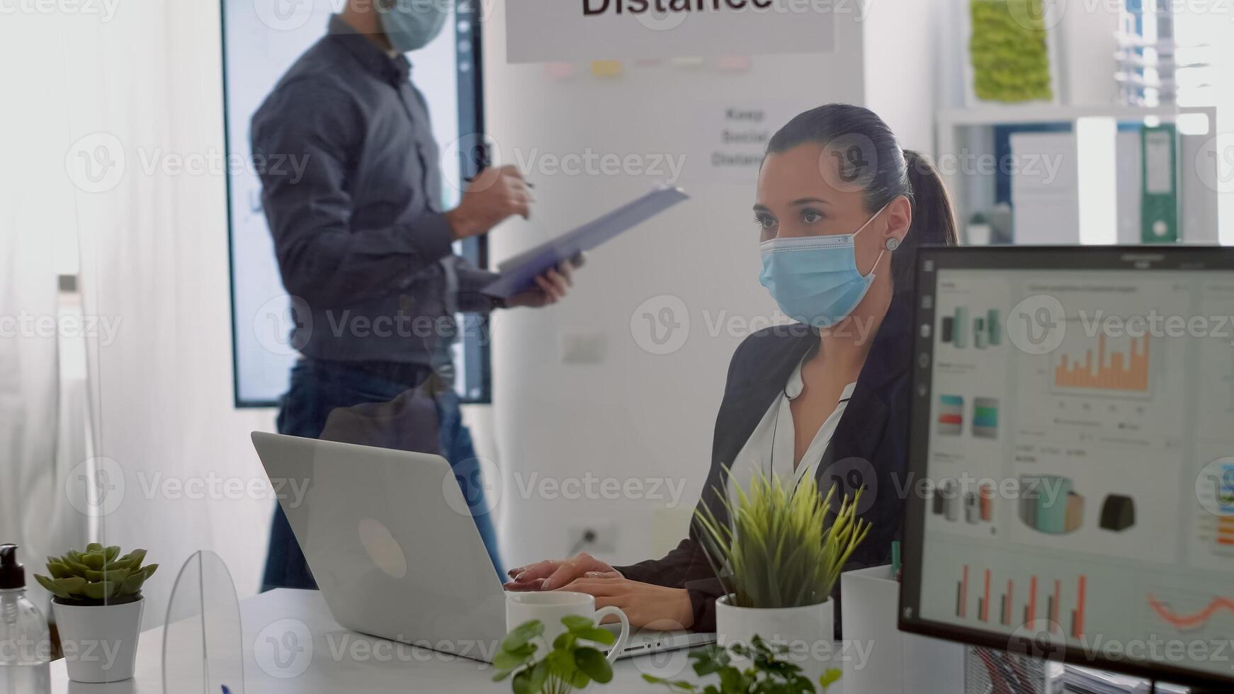 Executive manager with face mask typing marketing information on laptop computer while sitting at desk table in company office. Team respecting social distance to avoid infection with covid19 photo
