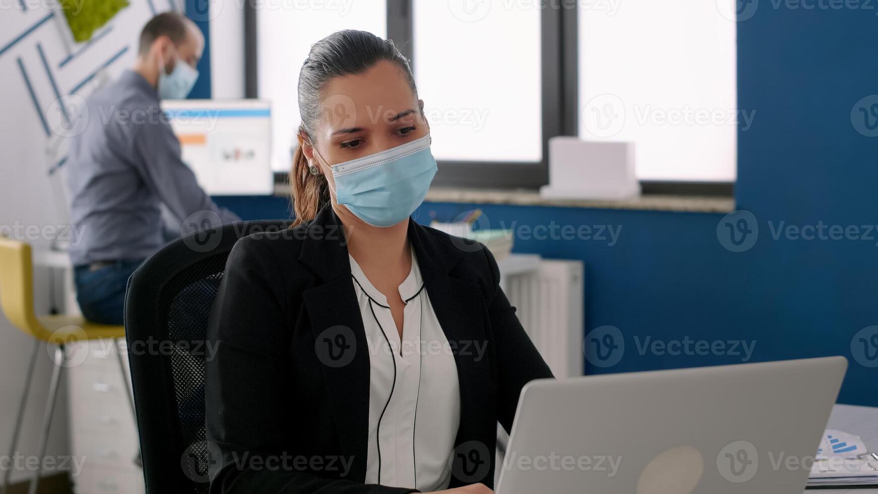 Business woman with protective face mask analysing on laptop computer marketing statistics, sitting at desk table in company office. Coworkers keeping social distancing to avoid virus disease photo
