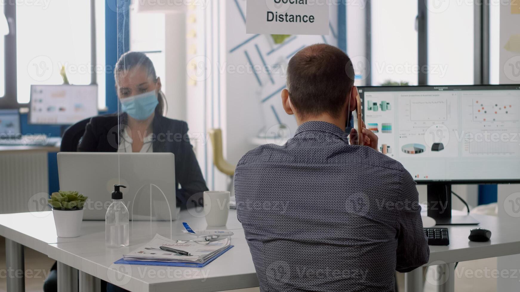 Business man wearing face mask talking at phone while sitting at desk in company office during coronavirus global pandemic. Coworkers keeping social distancing to avoid virus disease photo