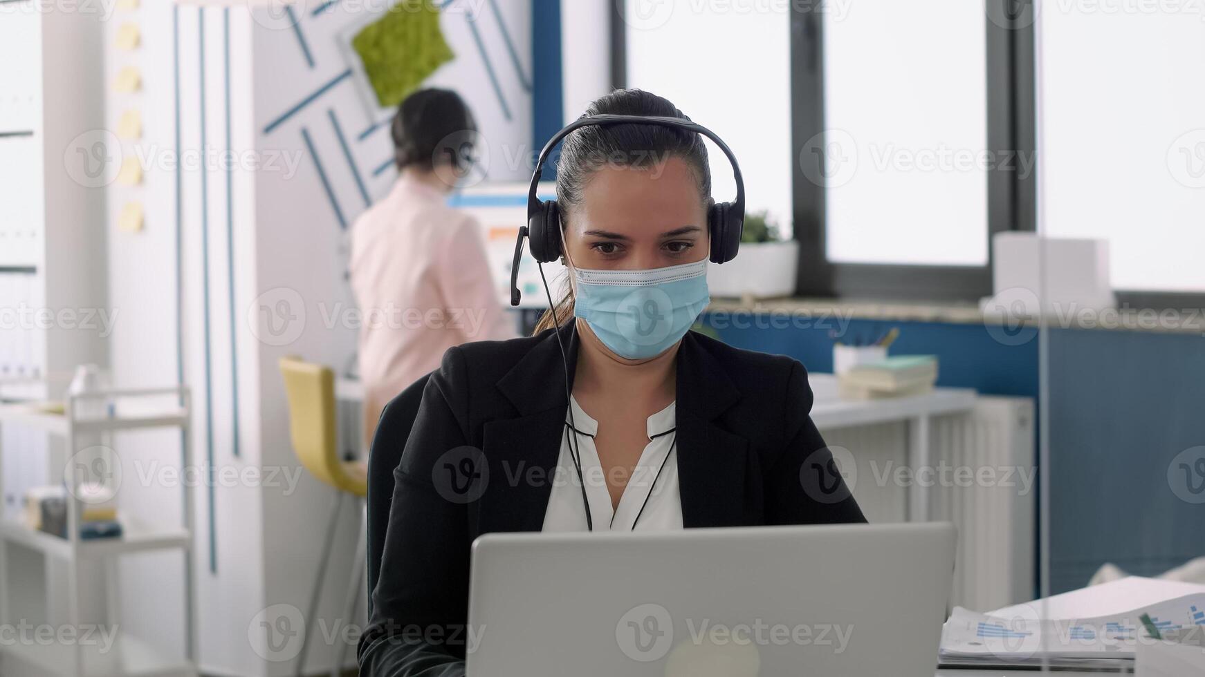 Businesswoman with protection face mask talking into microphone sitting at table desk in front of laptop computer. Coworkers keeping social distancing to prevent virus disease photo