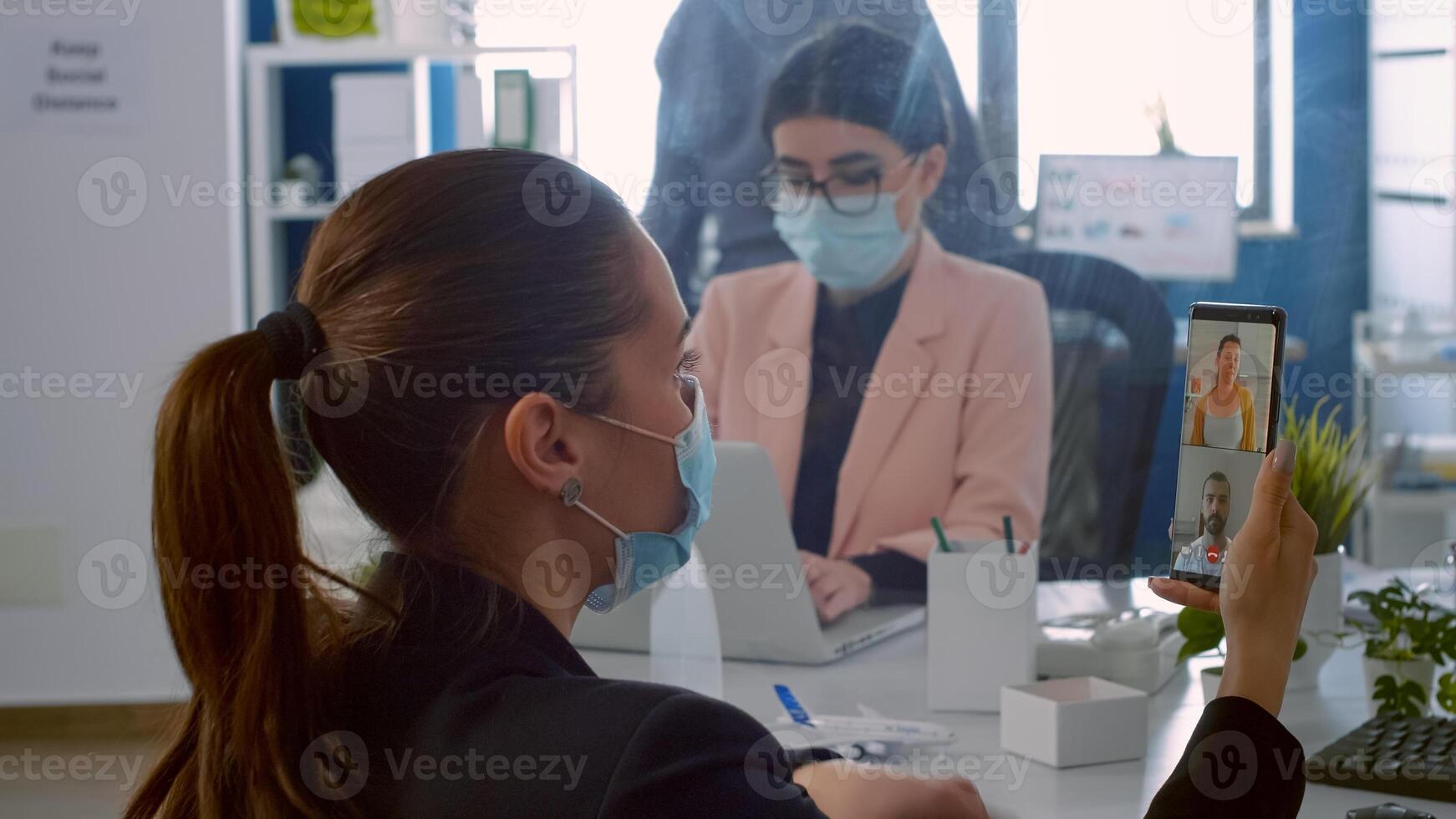 Back view of freelancer man with face mask talking on videocall meeting with remotely collegues using phone. Business woman working in new normal company office during covid19 pandemic photo