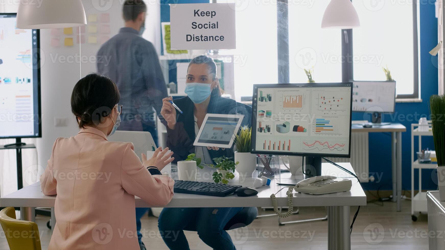 Executive manager with face mask showing statistics to her collegue using digital tablet computer. Coworkers working in new normal office keeping ing ing social distance to prevent infection with covid19 photo