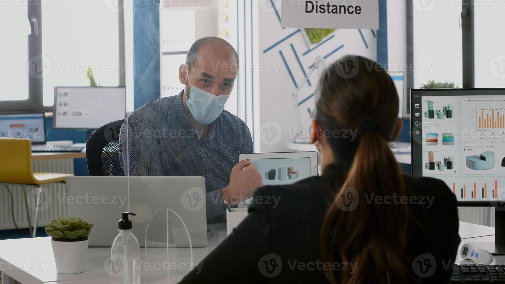 Coworkers with protective face masks talking about management statistics using tablet computer sitting in new normal company office. Team mantain social distancing to avoid infection with coronavirus photo