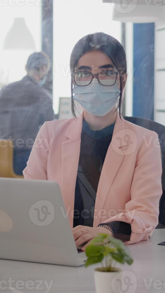 Portrait of business woman with protection face mask working on laptop computer in office during coronavirus pandemic. Coworkers keeping ing ing social distancing to prevent infection with covid19 virus photo