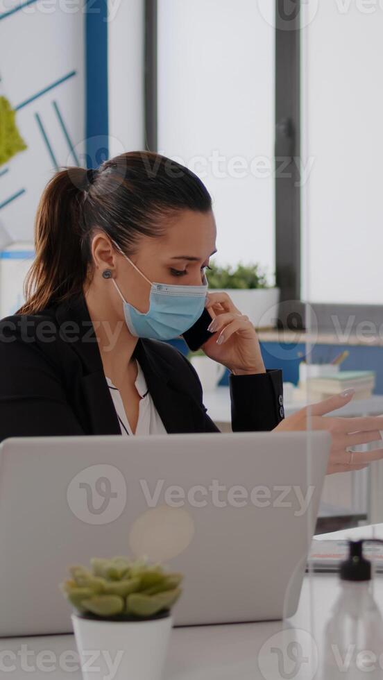 Woman with protective face mask working on laptop computer in company office checking statistics while talking on phone.Team workers respecting social distancing to avoid infection with covid19 virus photo