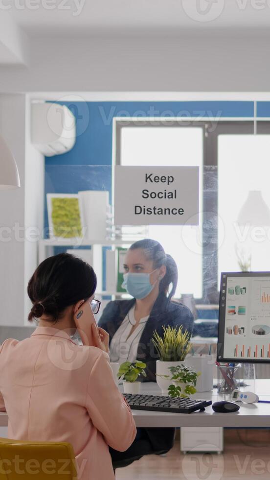 Business woman with medical face mask talking on smartphone with partners while her coworkers working in background respecting social distancing to prevent infection with coronavirus photo