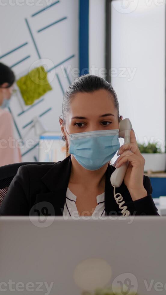 Entrepreneur with protective face mask discussing on landline while sitting at startup office desk in front of computer. Caucasian female working at business meeting during coronavirus global pandemic photo