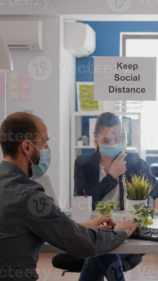 Freelancer with protection medical face mask working on laptop computer while talking on phone with team. Businesswoman sitting at desk table in new normal company office during covid19 quarantine photo
