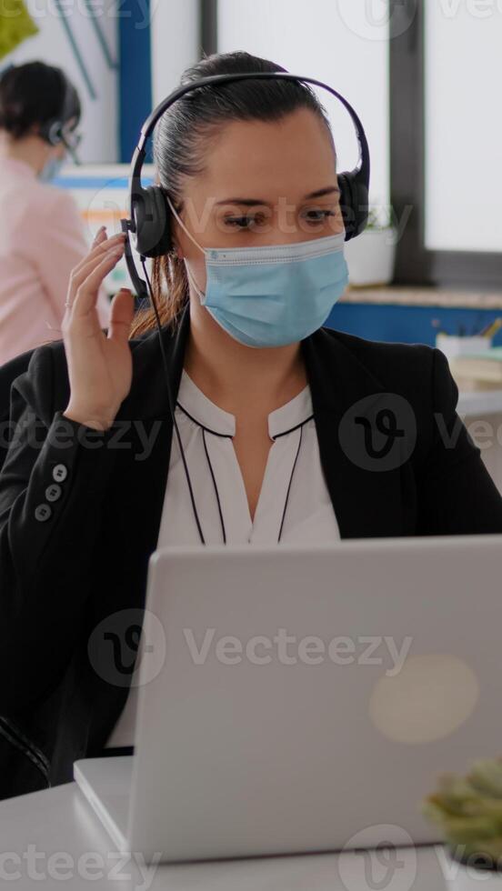 Close up of businesswoman with face mask wearing headset talking into microphone while typing marketing statistics on laptop computer. Freelancer sitting at desk in office during covid19 pandemic photo