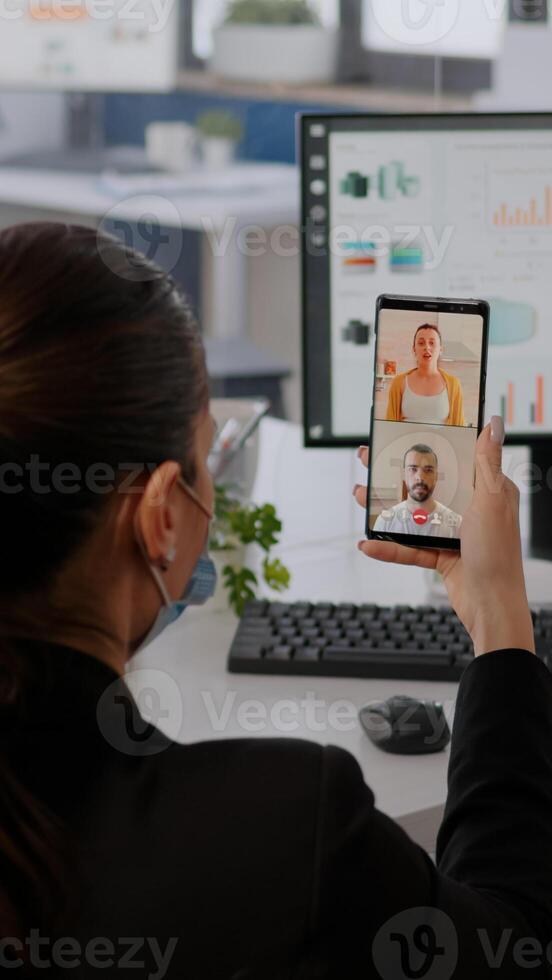 Executive business woman wearing protective face mask using phone for online videocall conference with remotely team. Coworkers working in background respecting social distance photo