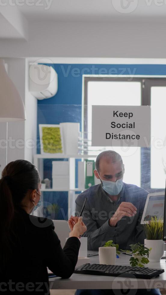 Business people working at financial graphs using digital tablet while sitting at office desk in corporate company. Team wearing face masks maintaining social distancing to avoid coronavirus pandemic photo