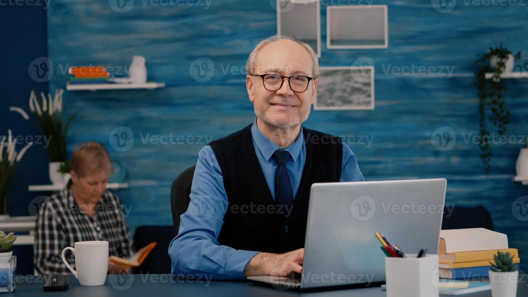 Retired manager sitting at desk in front of camera smiling after typing at laptop working from home while senior wife reading a book in background. Man prepared for online meeting using moden gadgets photo