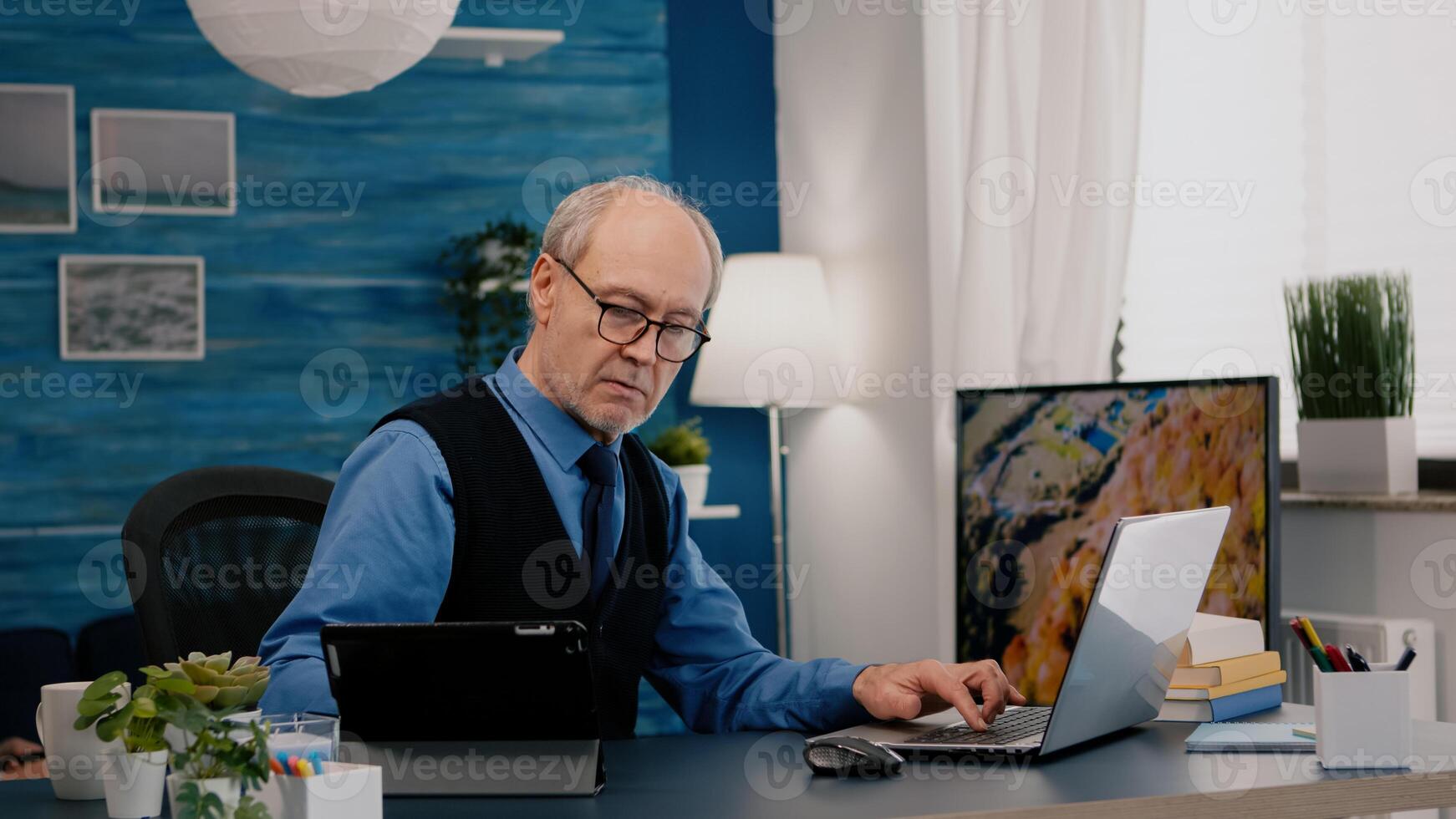 Senior man using laptop and tablet in same time working in office living room at project. Busy focused old employee writing, searching, analysing reports while elderly wife watching tv in background photo