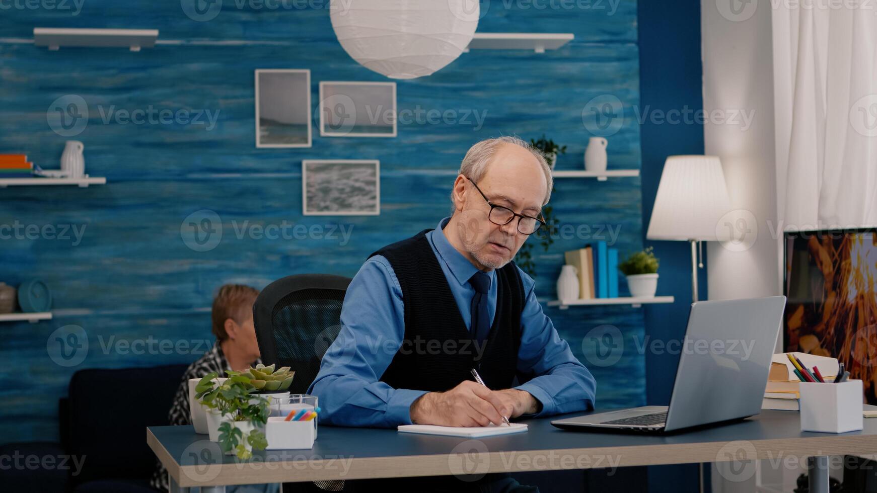 Multitasking retired man reading on laptop and writing on notebook working from home. Focused old remote employee searching, analysing financial reports while elderly wife watching tv in background photo