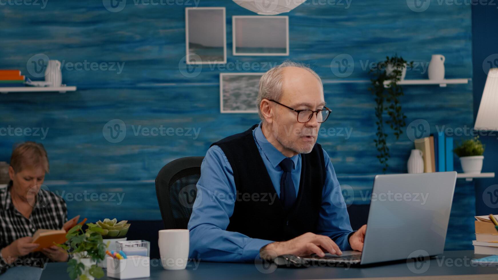 Old freelancer typing on laptop sitting in living room working remote. Busy focused elderly employee using modern technology reading typing, searching while old wife sitting on couch reading a book photo