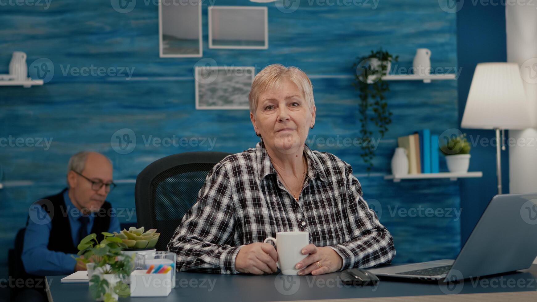 Senior focused woman looking at camera sitting in workplace at desk working from home while old wife watching tv in background. Retired business person prepared for online meeting using moden gadgets photo