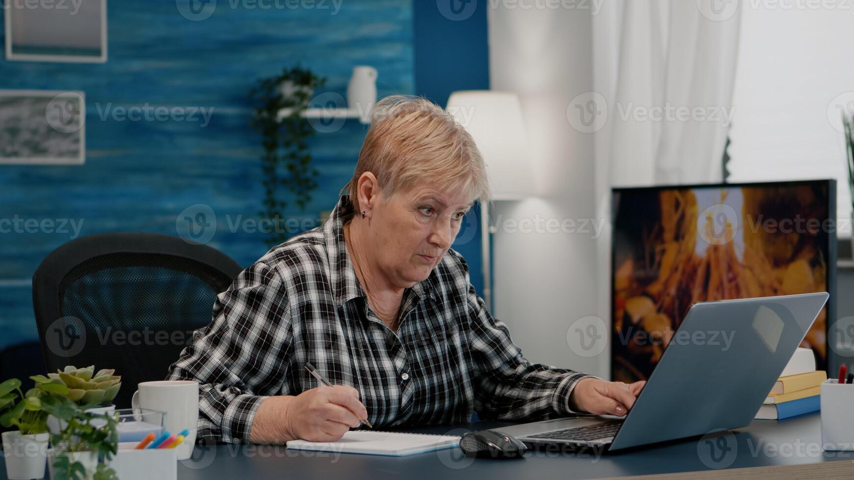 Old woman entrepreneur checking financial project of company looking on laptop analysing annual statistics data, taking notes on notebook sitting at desk in living room office working from home. photo