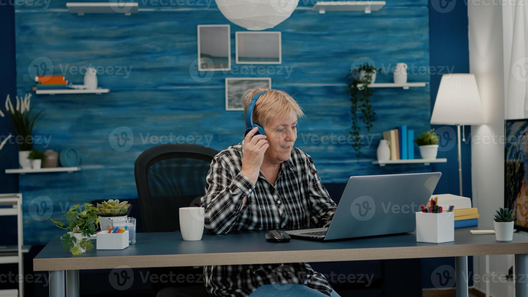 Happy old woman entrepreneur with headset listening music while checking statistics, writing on laptop working from home. Retired worker using modern technology reading typing, searching on computer photo