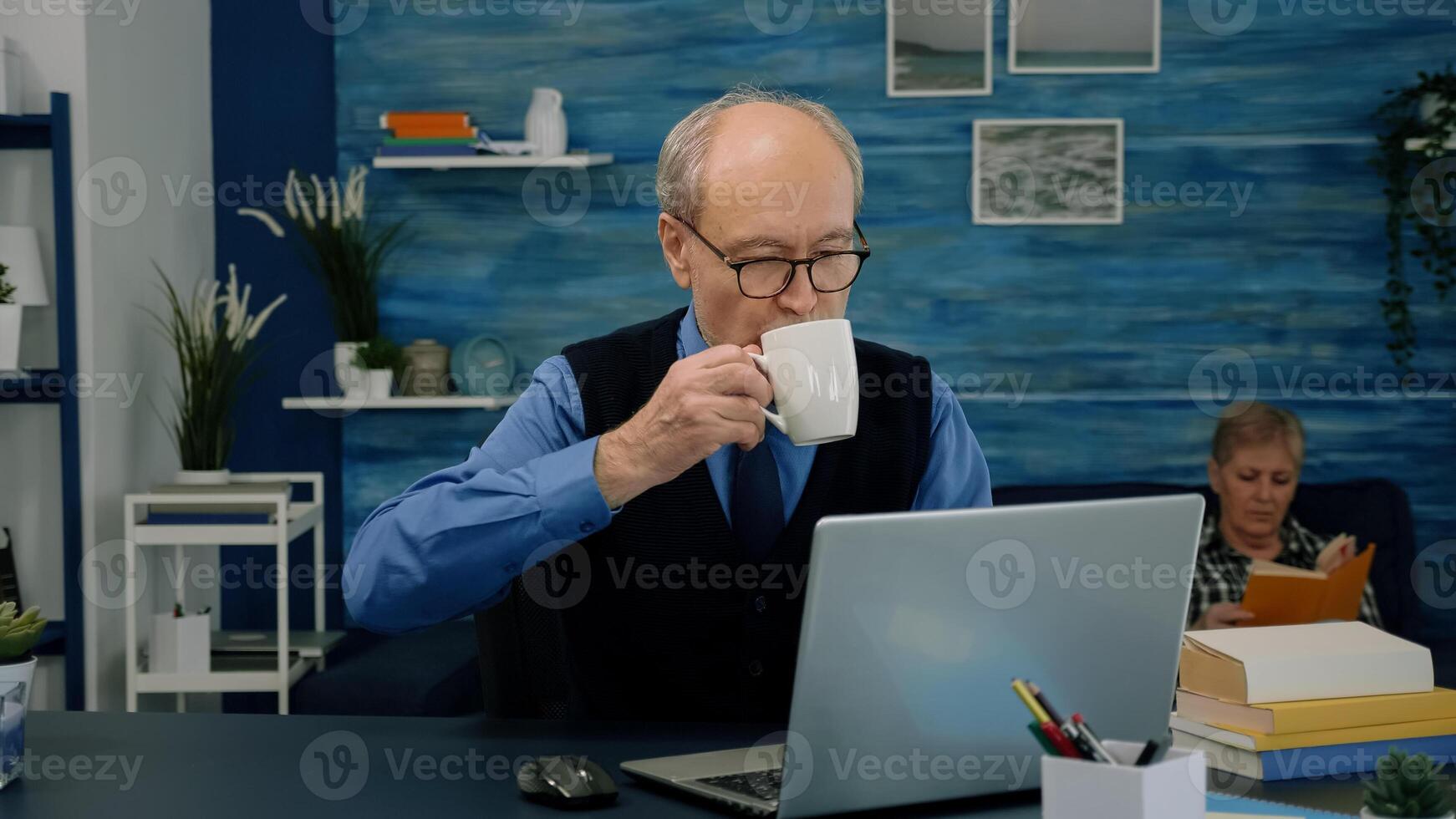 Tired elderly man sitting at desk and taking a sip of coffee, looking at laptop working from home workspace while his wife is reading a book in background. Focused old entrepreneur checking graphics photo