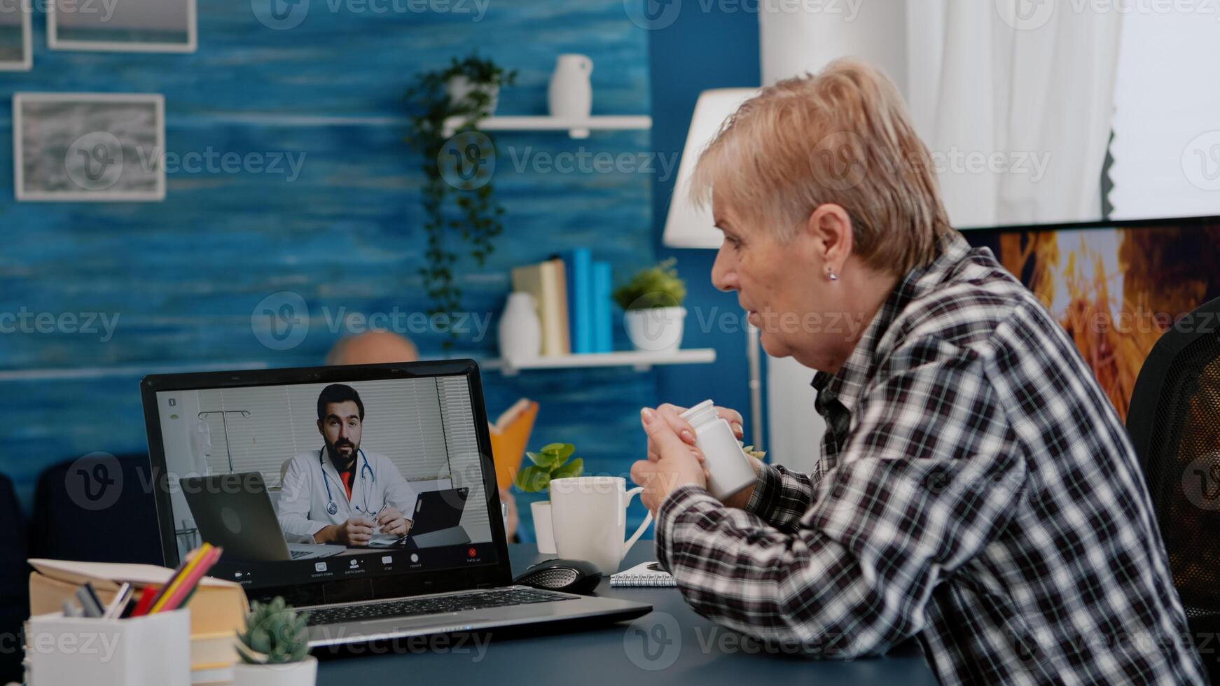 Old woman sitting at table at home looking at laptop answering question about health talks with therapist by videoconference call application, holding pills flask Remote medical consultation concept photo