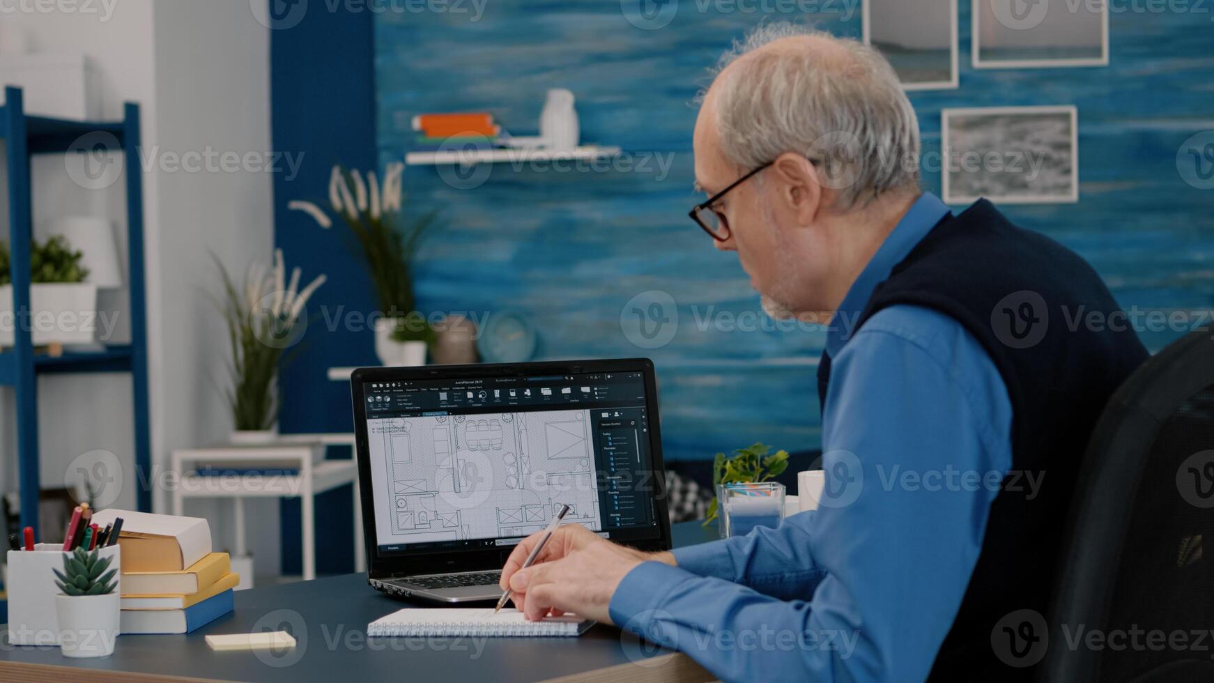 Senior man architect analysing digital prototype with plans from laptop working from home, taking notes on notebook. Elderly engineer designer using cad software to create a 3D concept of buildings. photo