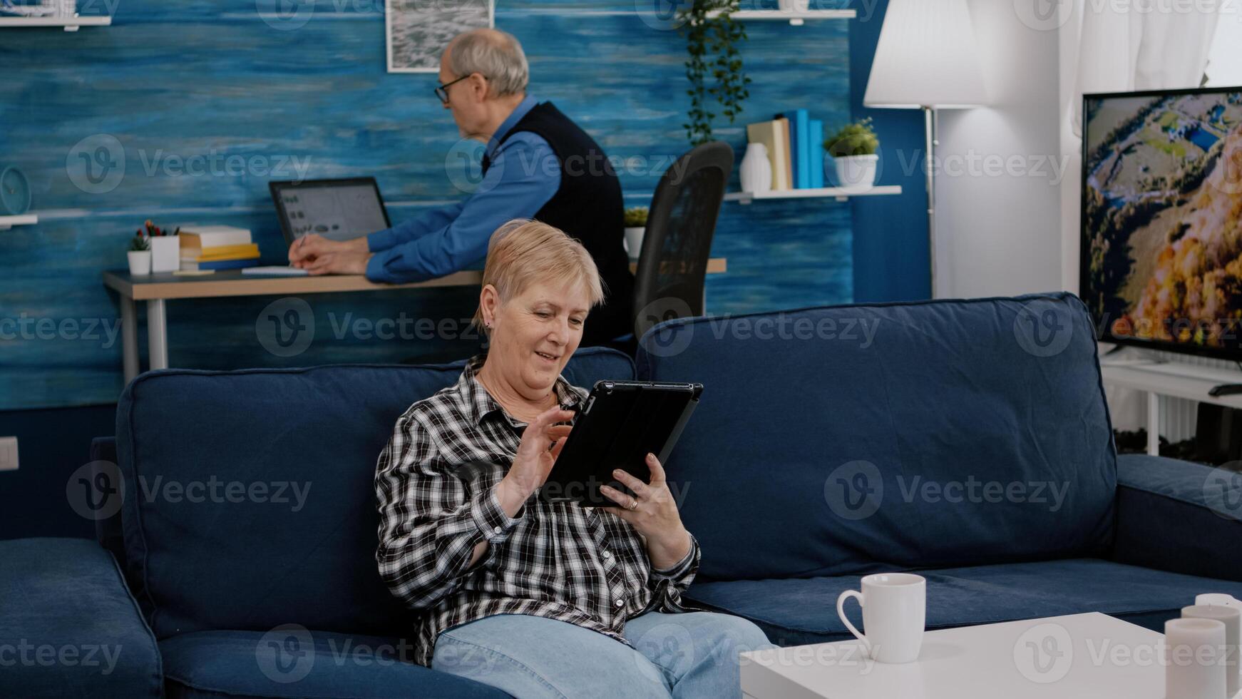 Happy old senior grandmother holding digital tablet browsing, searching informations, shopping online, while adult man working at laptop in background. Person enjoying using notepad gadget photo