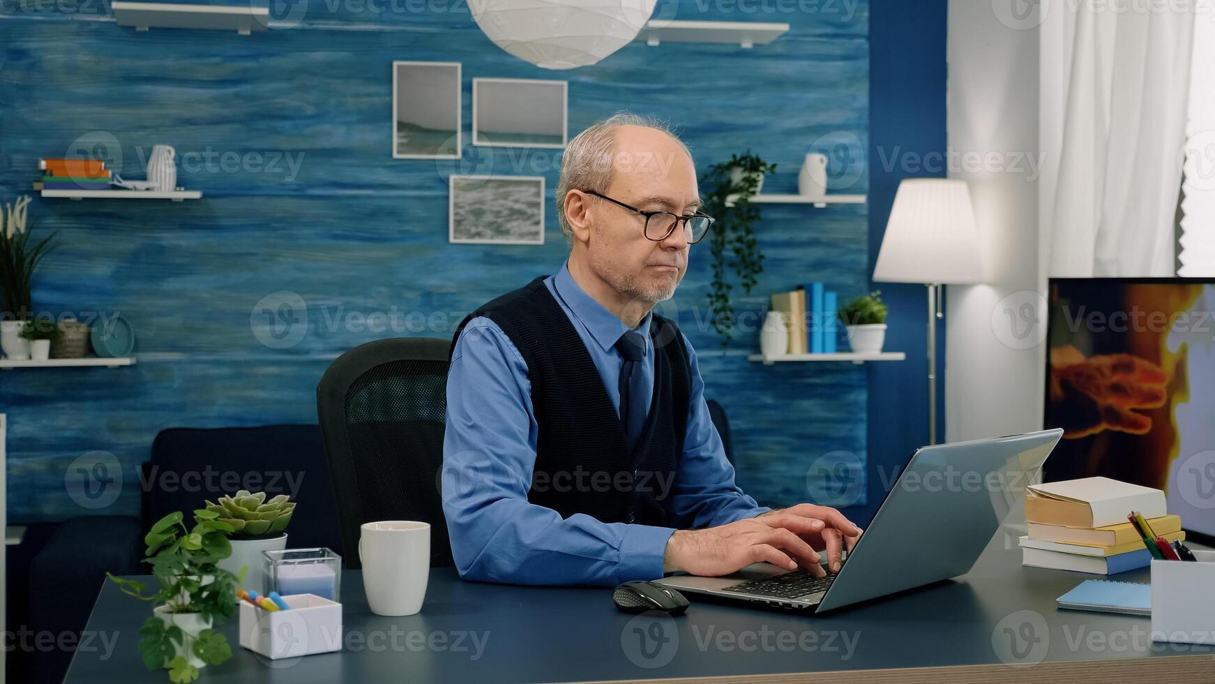 Senior remote businessman opening laptop and reading reports working from home drinking coffee. Retired man using modern technology analysing, searching info while wife sitting on couch reading a book photo