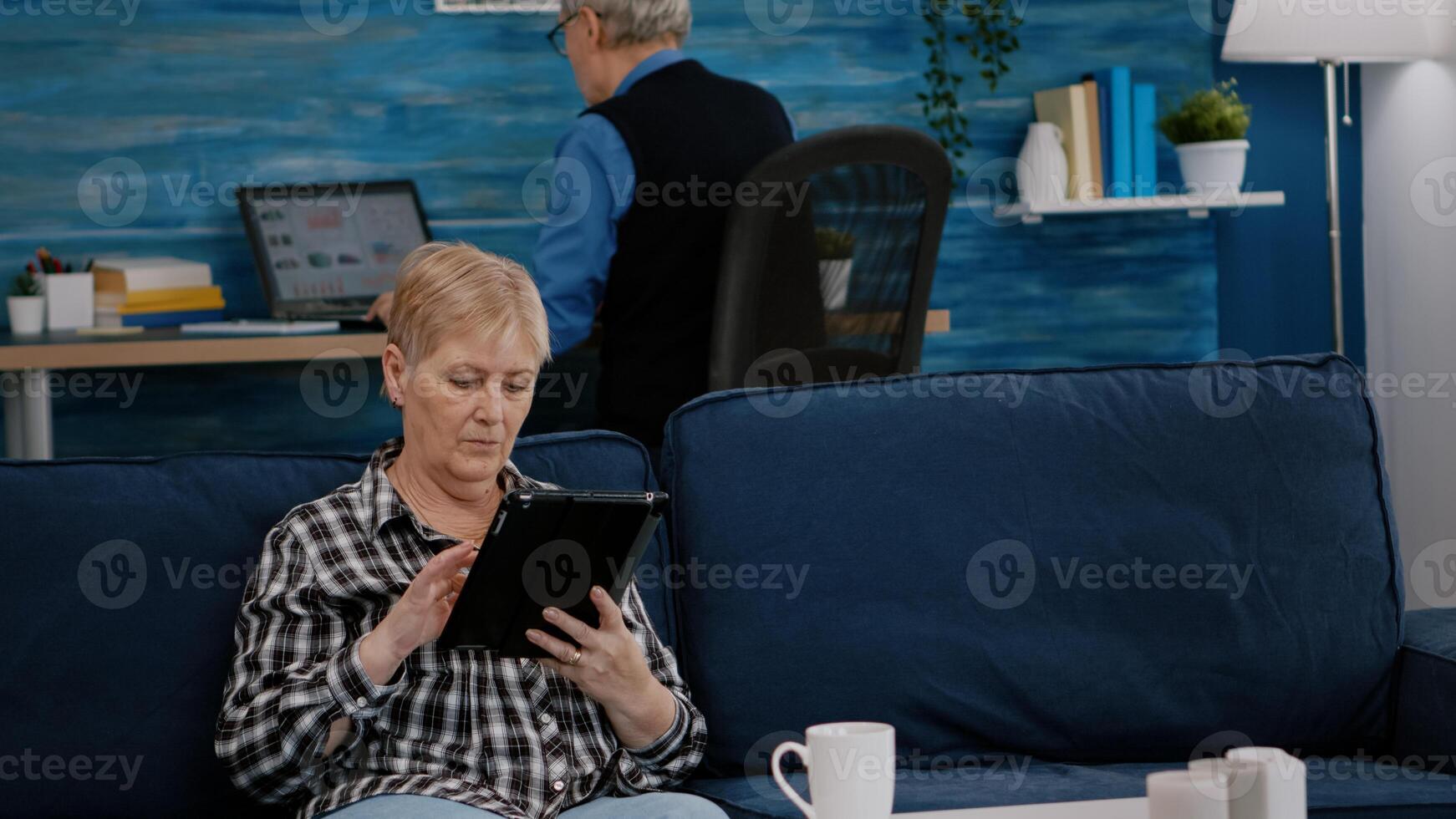 Middle aged old woman relaxing holding tablet reading e book sitting on sofa at home, while senior adult man working at laptop in background. Person enjoying using notepad browsing internet shopping photo