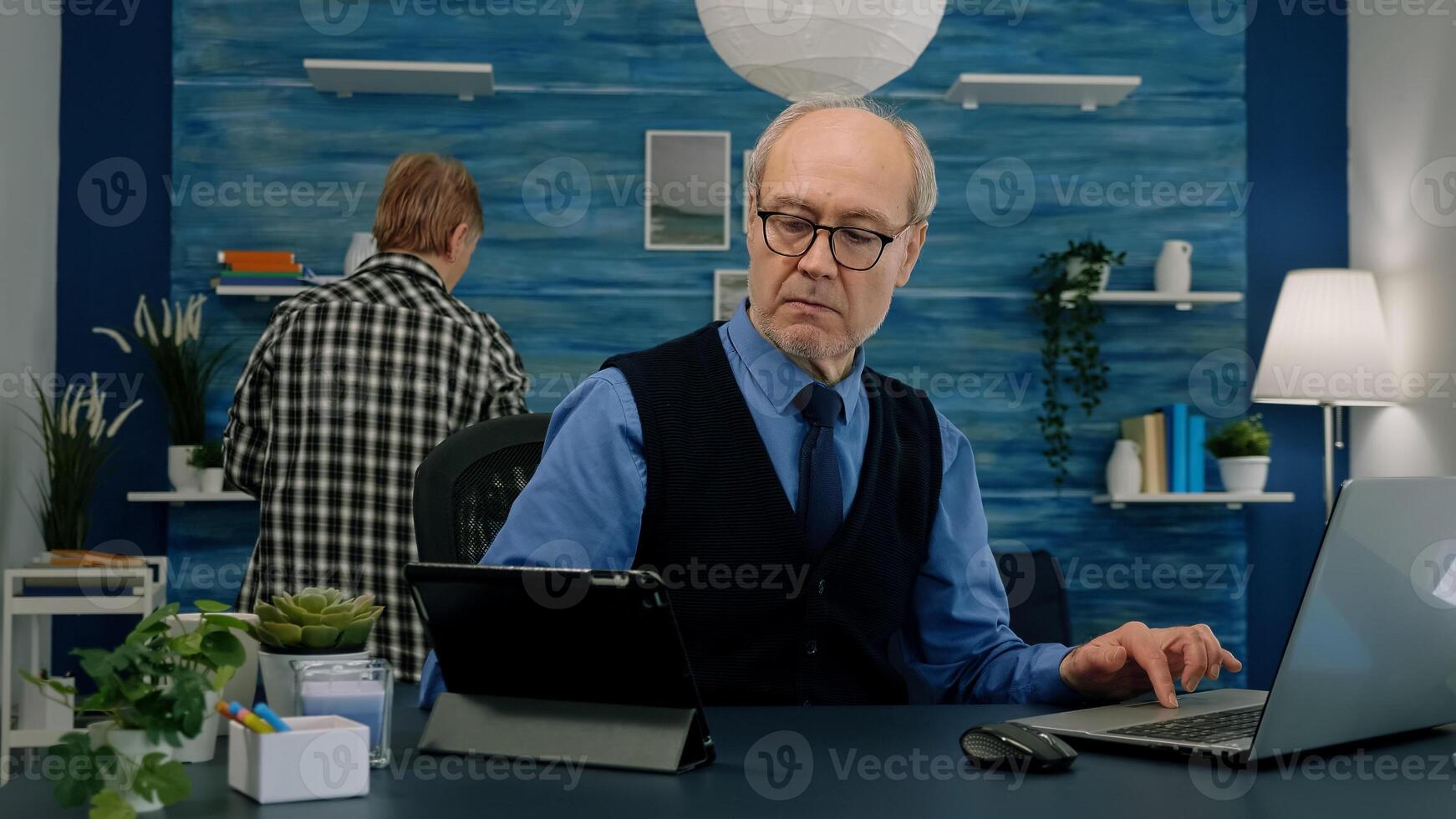 Elderly man using tablet and laptop in same time compering financial graphs working from home sitting in workplace. Busy focused old employee writing, searching, analysing reports photo