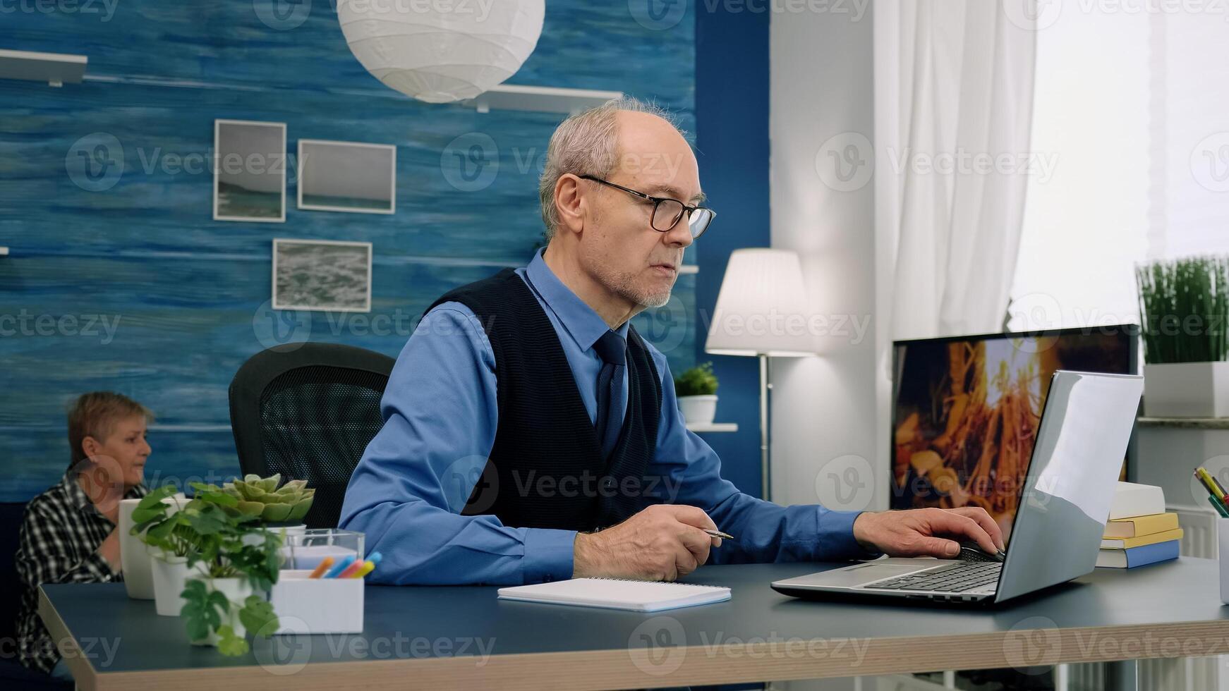 Middle age remote businessman working at laptop from home taking notes on notebook in modern living room. Busy focused employee using modern network technology reading writing, searching for job photo