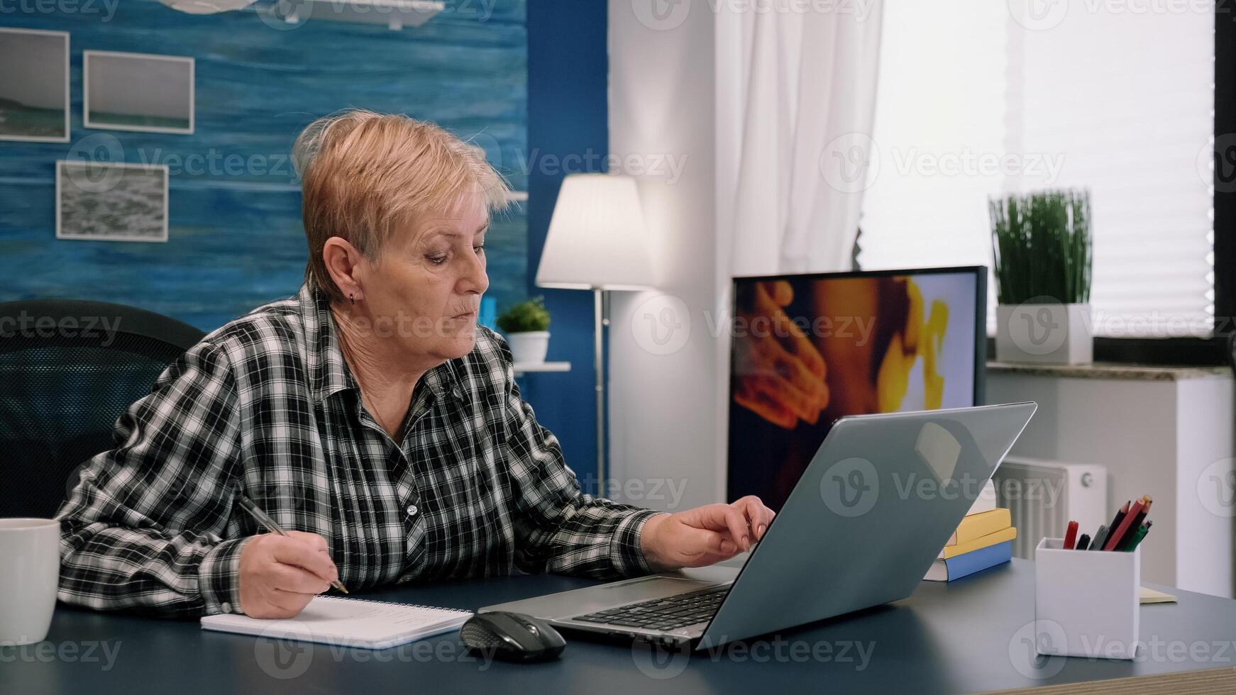Senior stylish woman taking notes in notebook while using laptop at home. Old freelancer writing details on book while working from workspace in living room, checking financial project of company photo