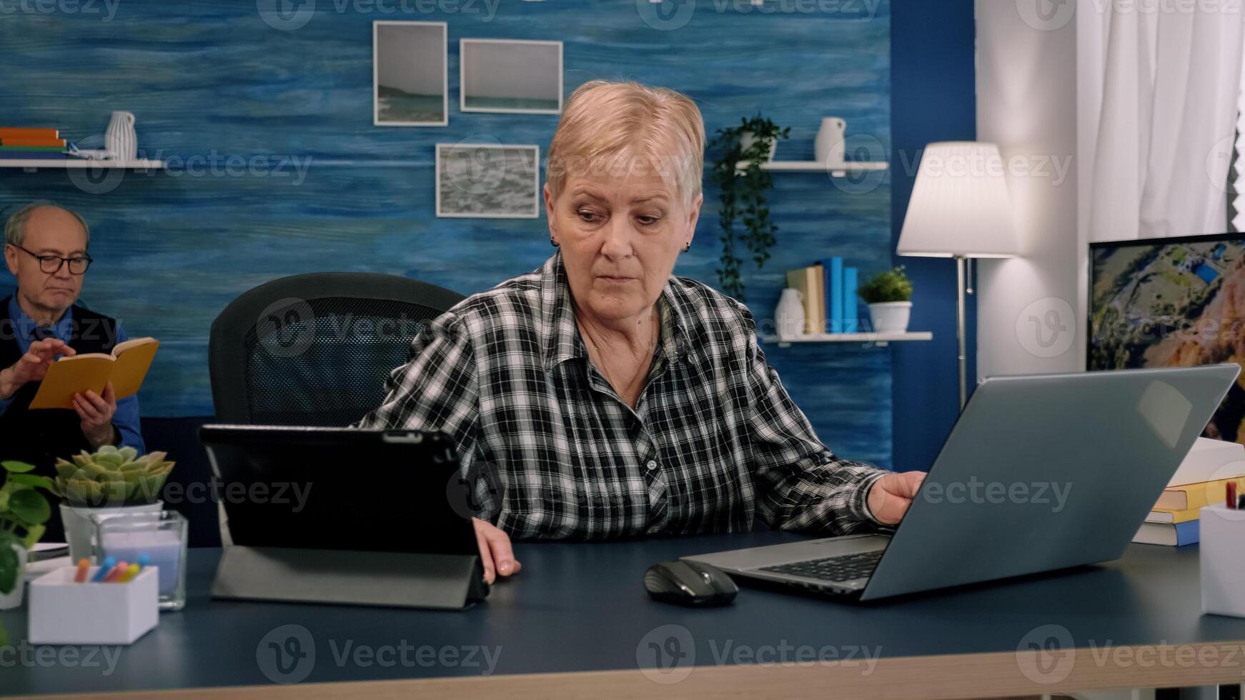 Old manager woman using laptop and tablet in same time working in home workspace for start-up company. Multitasking employee using modern technology network wireless overworking writing, searching photo
