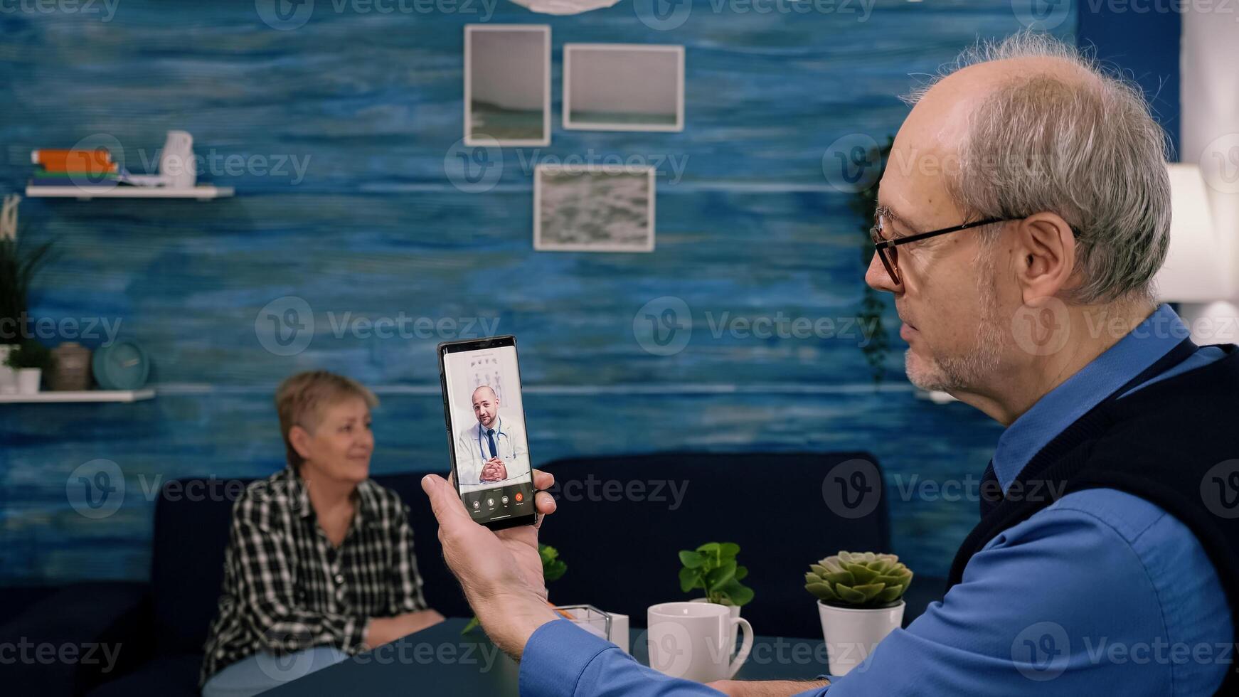 Retired male holding smartphone during online consultation listening young physician doctor sitting in living room. Sick man discussing during conference about symptoms, while wife looking at tv photo