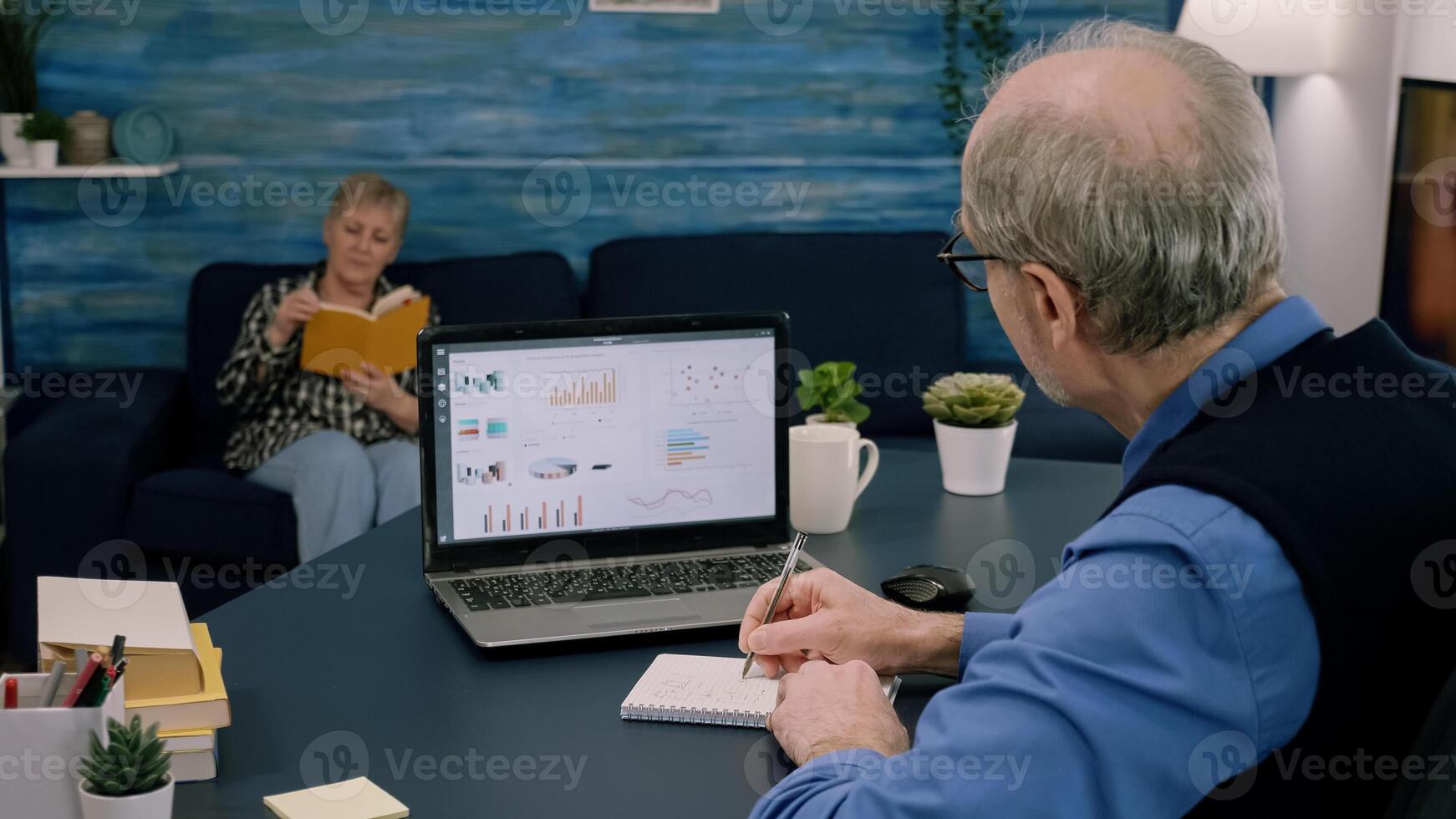 Tired elderly entrepreneur checking graphics from laptop writing on notebook working from business office overtime. Busy employee using modern technology network wireless reading typing, searching photo