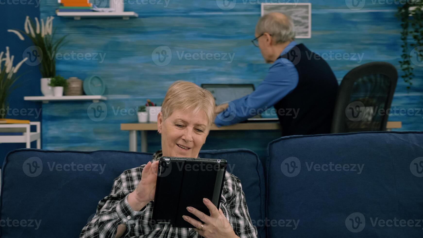 Senior woman reading emails on digital tablet sitting on sofa at home, while senior adult man working at laptop in background. Person enjoying using notepad browsing internet shopping photo
