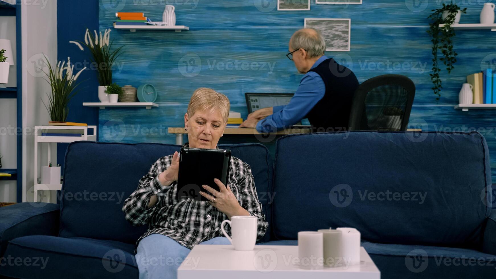 Focused senior grandmother relaxing sitting on sofa typing tablet browsing, searching informations, shopping online, while adult man working at laptop in background. Person using notepad gadget photo
