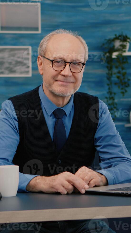 Senior man looking at camera smiling sitting in workplace at desk working from home while old wife watching tv in background. Retired business person prepared for online meeting using moden gadgets photo