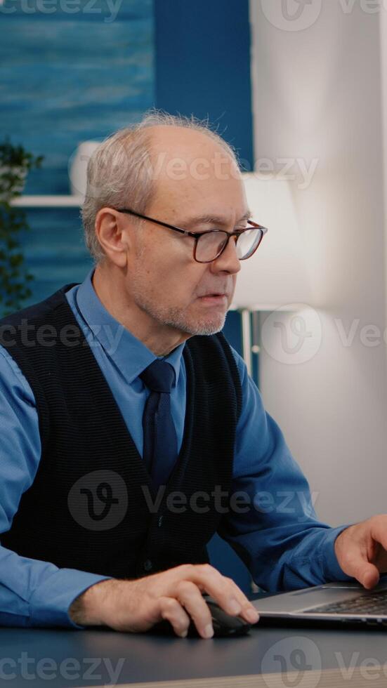 Focused old entrepreneur checking graphics writing on laptop working from home drinking coffee. Retired employee using modern technology reading typing, searching while senior wife sitting on couch photo