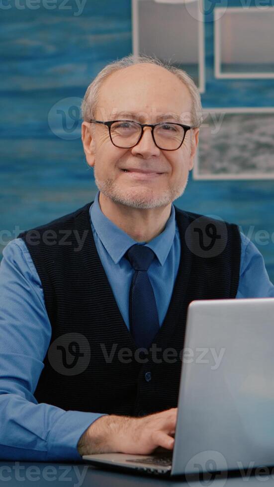 Retired manager sitting at desk in front of camera smiling after typing at laptop working from home while senior wife reading a book in background. Man prepared for online meeting using moden gadgets photo