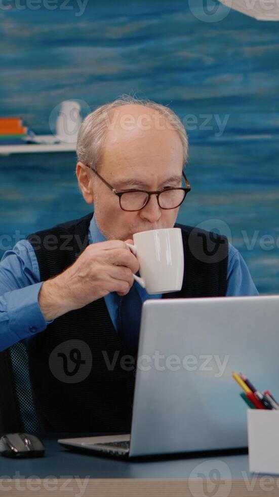 Senior businessman reading reports sitting in front of laptop working from home drinking coffee. Retired man using modern technology analysing typing searching while wife sitting on couch reading a book photo