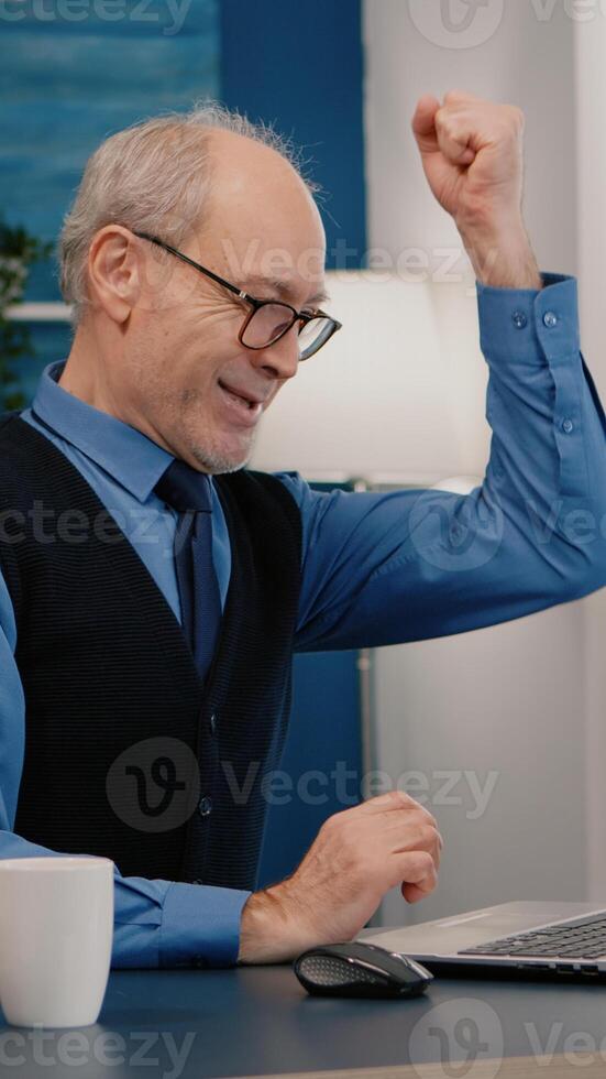 Excited senior man feel ecstatic reading great online news on laptop working from home. Retired employee using modern technology reading typing, searching while senior wife sitting on couch photo