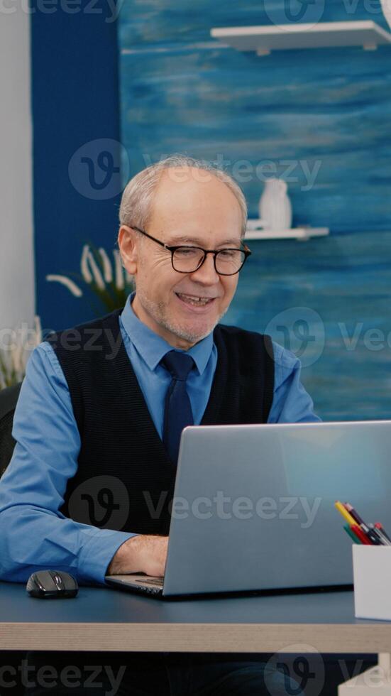 Happy old manager receving good news on laptop working from home for startup company, gesturing victory. Retired employee using modern technology reading typing searching while senior wife watching TV photo