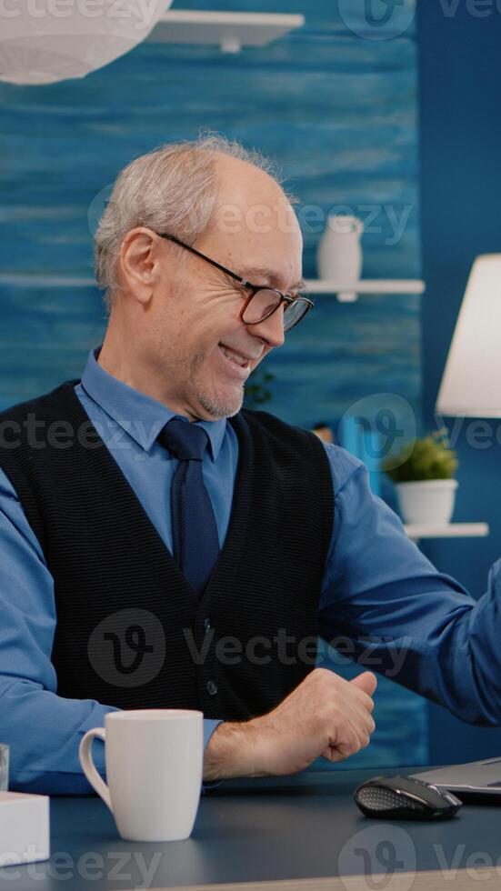 Old freelancer receving good news on laptop working from home sitting in living room. Enthusiastic senior employee using modern technology reading typing, searching while senior wife sitting on couch photo